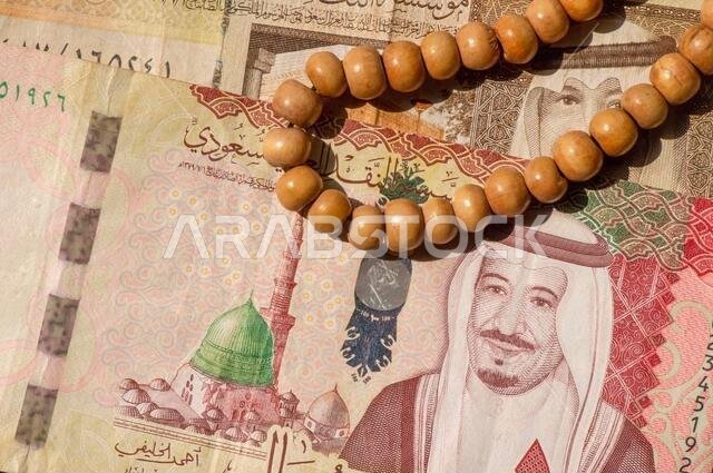 صورة علوية للنقود، الأوراق النقدية والعملات المعدنية للريال السعودي، ومسبحة الصلاة، بتركيز ضحل.