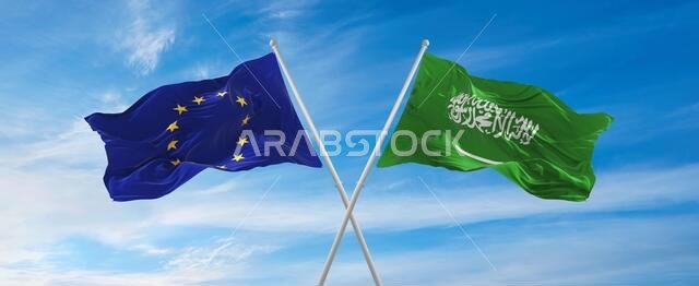 أعلام الاتحاد الأوروبي والمملكة العربية السعودية ترفرف في مهب الريح على أعمدة الأعلام في سماء ملبدة بالغيوم في يوم مشمس. ترمز إلى العلاقة والحوار بين