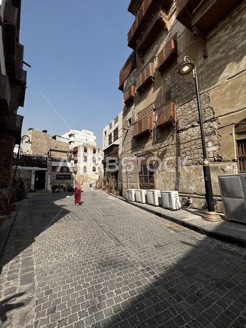 صورة من الخلف لامرأة عربي خليجية سعودية تتجول في حواري البلد التاريخي في جدة، المباني الأثرية في البلد التاريخي، التصميم المعماري العربي القديم، المعالم التاريخية في جدة، السياحة في السعودية