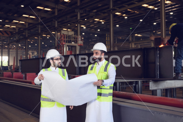 Two Saudi Arabian Gulf industrial engineers wearing a protective jacket and helmet are supervising the production in the factory, holding the factory blueprint sheet, the steps of the production process, the concept of engineering and industry