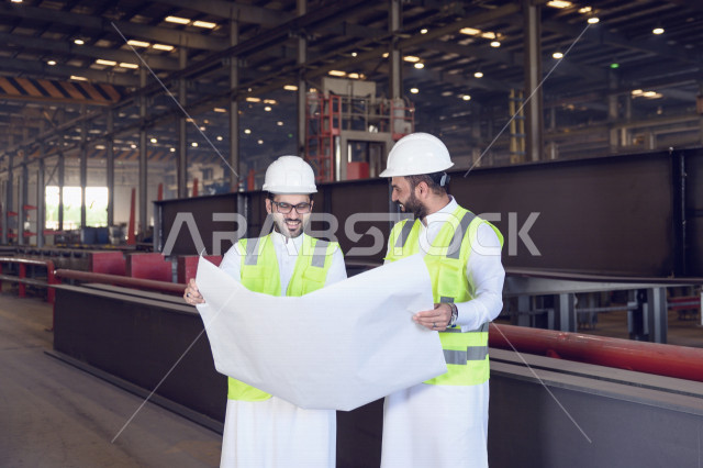 مهندسان صناعيان عربيان خليجيان سعوديان يرتديان سترة و خوذة الوقاية و الحماية  يشرفان على الانتاج في المصنع ، يحملان ورقة مخطط المصنع  ، خطوات العملية الانتاجية ، مفهوم الهندسة و الصناعة
