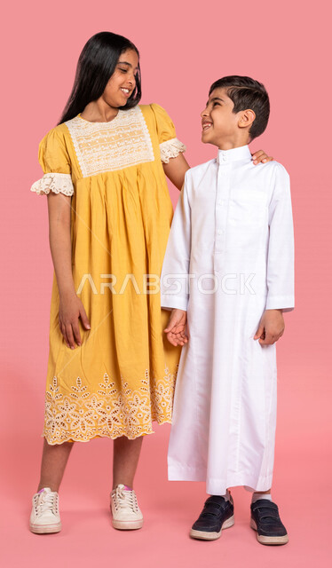 ورتريه لطفلان عربيان خليجيان سعوديان، العلاقات الأخوية المترابطة، علاقات اجتماعية، تعارف وصداقات، إيماءات وجه ويدين مختلفة تدل على السعادة، الوقوف بثقة، صورة الجسم بالطول الكامل، خلفية ملونة