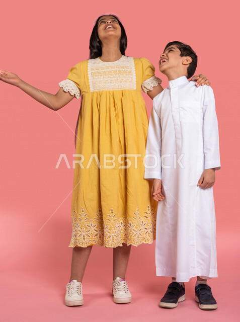 ورتريه لطفلان عربيان خليجيان سعوديان، العلاقات الأخوية المترابطة، علاقات اجتماعية، تعارف وصداقات، إيماءات وجه ويدين مختلفة تدل على السعادة، الوقوف بثقة، صورة الجسم بالطول الكامل، خلفية ملونة