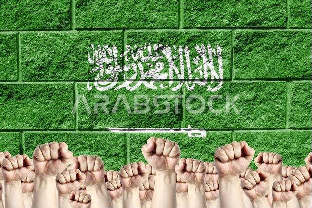 Raised fists against the background of the depicted flag of Saudi Arabia on a brick wall, a concept of strength and unity.