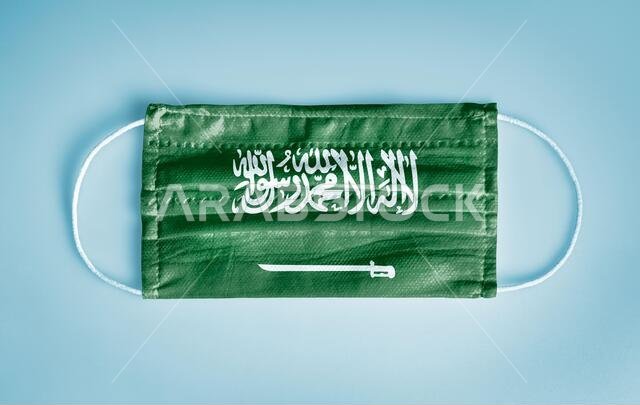 Covid-19 Coronavirus protection concept: Medical disposable face mask with Saudi Arabia flag on blue background.