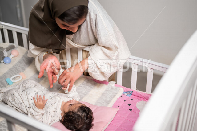 أم عربية خليجية سعودية مع طفلها الرضيع مستلقي في المهد، تلاعب طفلها بإيماءات تدل على السعادة والامان، مفهوم الأمومة والطفولة، حب وحنان الأمهات، رعاية الأم لأطفالها والعناية بهم، قضاء أوقات جميلة مع الاطفال، الأسرة وتحمل المسؤولية