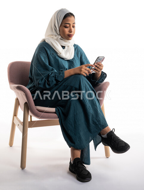 بورتريه لإمرأة عربية خليجية سعودية تجلس على الكرسي وتستخدم الهاتف المحمول ، استخدام البرامج والتطبيقات الالكترونية، التسوق الالكتروني، التواصل الاجتماعي، تصفح المواقع الالكترونية، ادمان الانترنت، خلفية بيضاء