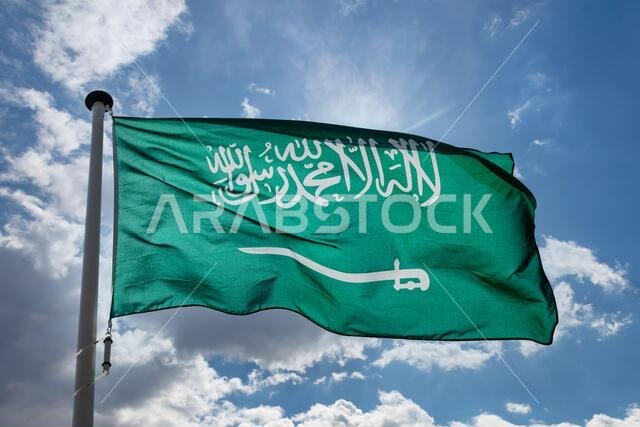 علم المملكة العربية السعودية، رمز المملكة العربية السعودية الوطني، يرفرف على سارية علم في سماء زرقاء ملبدة بالغيوم