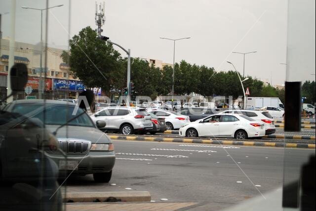 الرياض، المملكة العربية السعودية - ١٨ فبراير ٢٠٢١: حركة مرور على الطريق بانتظار الإشارة، أشجار ومتاجر في الخلف، طقس غائم جميل