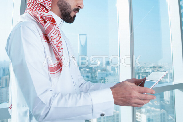 صورة مقربة لرجل اعمال عربي خليجي سعودي يستخدم جهاز تقني التابلت ، تصفح الإنترنت ، جلسة عمل ، العمل عن بعد ، خلفية لأبراج الرياض ، عقد إجتماع عمل عبر الإنترنت ، التسويق الإلكتروني ، إستخدام التكنولوجيا وأجهزة تقنية حديثة