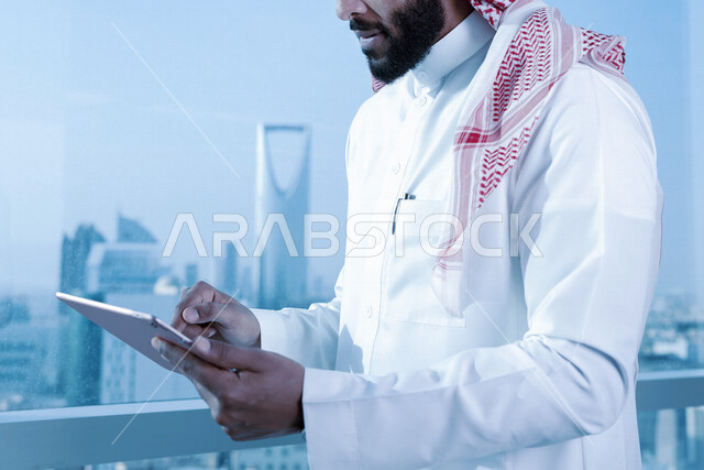 صورة مقربة لرجل اعمال عربي خليجي سعودي يستخدم جهاز تقني التابلت ، تصفح الإنترنت ، جلسة عمل ، العمل عن بعد ، خلفية لأبراج الرياض ، عقد إجتماع عمل عبر الإنترنت ، التسويق الإلكتروني ، إستخدام التكنولوجيا وأجهزة تقنية حديثة