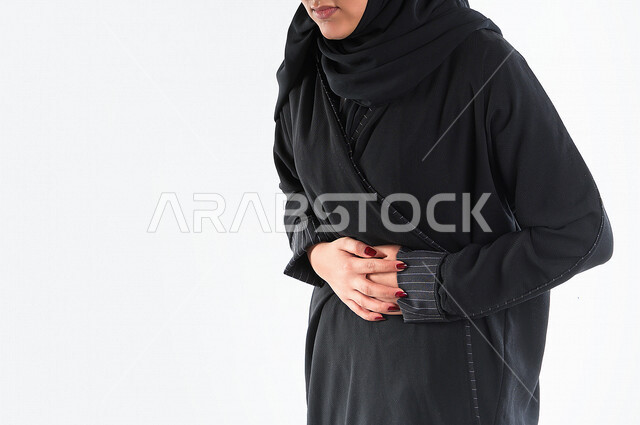 صورة مقربة لمرأه عربيه سعوديه خليجيه ترتدي العباءة والحجاب , إيماءات وجه ويدين تدل على الشعور بألم في البطن, الإحساس بالمغص, التعب والإرهاق, الحاجه للعلاج, طلب المساعده , خلفية بيضاء .