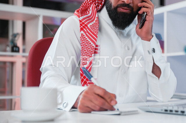 صورة مقربة لرجل عربي خليجي سعودي يقوم بإجراءمكالمة هاتفية وتدوين الملاحظات ، جلسة عمل ، متابعة المشاريع والمهام ، مهن وأعمال تجارية ، متابعة ومناقشة العمل ، عقد إجتماع عمل عبر الإنترنت