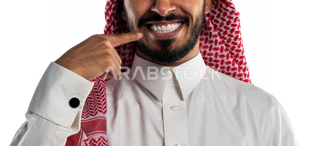 بورتريه بصورة مقربة لرجل عربي سعودي خليجي يشير بإًصبعه السبابة على أسنانه، يبتسم إبتسامة هوليود، استخدام قشور فينير من الخزف للأسنان، تجميل وتبييض الاسنان، معالجة الأسنان، خلفية بيضاء، أسنان نظيفة مثالية