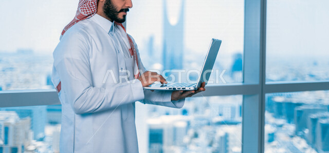 صورة مقربة لرجل اعمال عربي خليجي سعودي يستخدم جهازالحاسوب المحمول، تصفح الإنترنت، جلسة عمل، العمل عن بعد، خلفية لأبراج الرياض، عقد إجتماع عمل عبر الإنترنت، التسويق الإلكتروني، إستخدام التكنولوجيا وأجهزة تقنية حديثة
