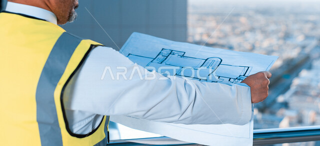 صورة مقربة بزاوية من الخلف لمهندس معماري عربي خليجي سعودي يرتدي سترة الوقاية الخاصة بالعمل ، يحمل بيده مخطط البناء ، متابعة المشروع وتدقيق العمل ، الاشراف والرقابة على المشاريع الهندسية ، الهندسة المعمارية ، مهن ووظائف هندسية