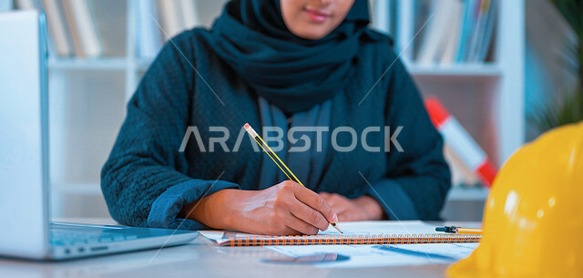 مهندسة عربية خليجية سعودية محجبة في مقر الشركة الهندسية، تقوم بتدوين الملاحظات ، إستخدام جهاز الحاسوب المحمول، مفهوم الهندسة المعمارية، إدارة المشاريع الهندسية، مهن ووظائف هندسية، خوذة وقاية، اشراف ومتابعة العمل، مخططات تصاميم هندسية