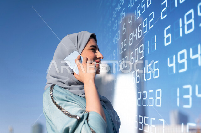 صورة مقربة لمرأة عربية خليجية سعودية محجبة، تقوم بإجراء مكالمة هاتفية ، إحصائية للبورصة المالية بتقنية الهولوجرام ثلاثية الأبعاد ، الإستثمار المالي ، مراقبة الإقتصاد والعملات الرقمية ، متابعة سوق الأوراق المالية