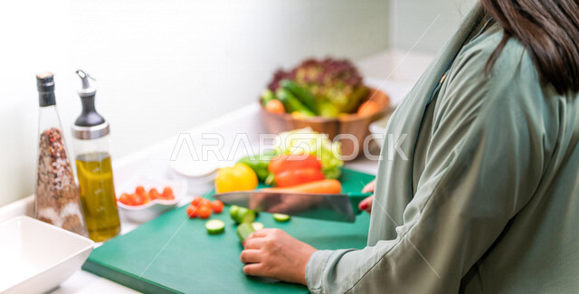 صورة مقربة بزاوية من الخلف لمرأة عربية خليجية سعودية، تقف أمام طاولة عليها مجموعة من الخضراوات الطازجة، إعداد السلطات والأطباق الصحية، مفهوم النظام الغذائي الصحي، تحضير وجبات طعام صحية ، فطور الصحي، سلطة خضار
