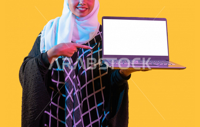بورتريه بصورة مقربة لمرأة خليجية سعودية محجبة، إستخدام جهاز الحاسوب المحمول، عرض شاشة لابتوب بيضاء مفرغة، جلسة عمل، تصفح الإنترنت، عرض خدمات وتطبيقات عبر اللابتوب، التسويق الرقمي الإلكتروني، مفهوم الإعلانات، خلفية صفراء