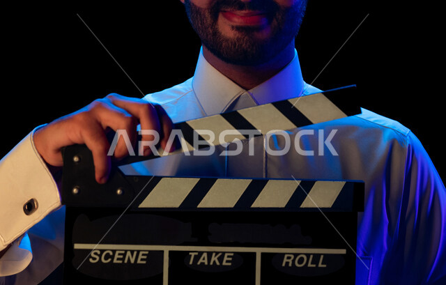 بورتريه بصورة مقربة لرجل عربي سعودي خليجي داخل موقع التصوير يحمل بيده لوح تصوير ، إخراج المسلسلات والأفلام ، ادارة العمل الفني ، الاشراف على طاقم التمثيل والعمل ، متابعة السيناريو والحوار ، التصوير السينيمائي ، خلفية سوداء