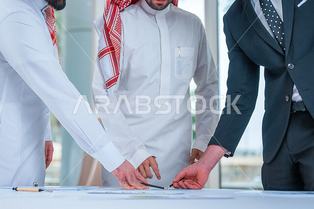 صورة مقربة لرجلان عربيان خليجيان سعوديان وبجانبهم رجل بالبدلة الرسمية يقومان بمتابعة ومناقشة الأعمال داخل مقر الشركة، مهن وأعمال تجارية، شركة سعودية، بيئة عمل، زملاء عمل، إدارة أعمال الشركة، تطوير المشاريع، عقد صفقات تجارية