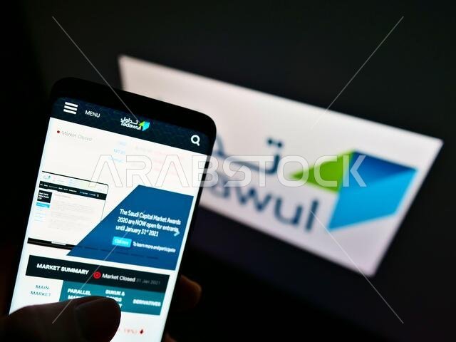 شخص يحمل هاتفًا ذكيًا يعرض موقع بورصة تداول السعودية على الشاشة أمام الشعار. التركيز على أعلى يسار شاشة الهاتف.
