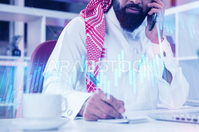 صورة مقربة لرجل عربي خليجي سعودي يجلس امام اللابتوب ويجري اتصال من خلال الهاتف المحمول، تسجيل وتدوين الملاحظات، اعداد التقارير المالية، تحليل البيانات المالية، رسومات بيانية لصعود وهبوط البورصة بتقنية الهولوجرام ثلاثية الأبعاد،