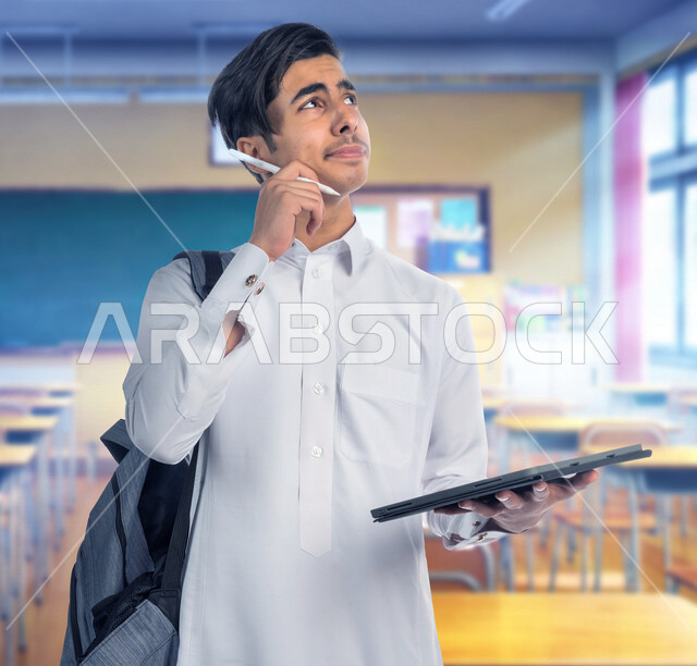 طالب عربي خليجي سعودي يرتدي يحمل بيده جهاز التابلت، كتابة وتدوين الملاحظات، متابعة المحاضرات الجامعية الالكترونية، حل الواجبات الالكترونية، التعليم الالكتروني، التعليم في المملكة العربية السعودية، ايماءات وجه ويدين تدل على التفكير بعمق، خلفية لفصل دراسي