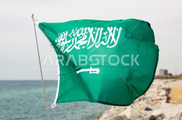 علم المملكة العربية السعودية يرفرف في مهب الريح على شاطئ البحر.