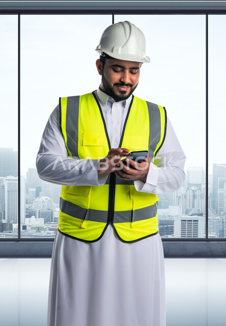 مهندس معماري عربي خليجي سعودي يرتدي السترة الواقية و خوذة السلامة ، يستخدم الهاتف المحمول ليتابع ادارة اعمال البناء  ، اشراف ومتابعة المشاريع,  تصفح الانترنت, الهندسه المعماريه