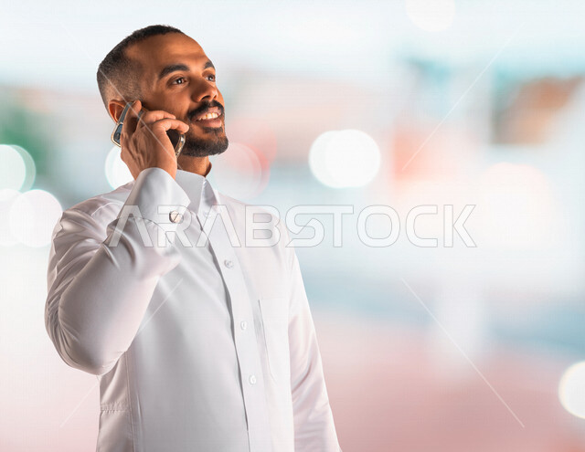 رجل عربي خليجي سعودي يرتدي الزي السعودي يقوم بإجراء مكالمة عبر الهاتف المحمول، دردشة مع الأصدقاء ، اجراء مكالمة عمل , إدارة الأعمال عن بعد، استخدام التقنية الحديثة .