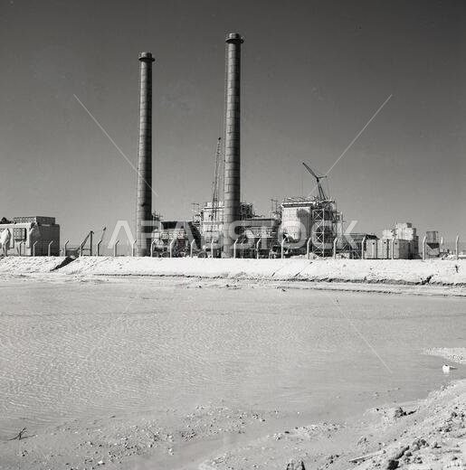 1960s, historical, Saudi Arabia, a small petro-chemical plant out in the desert, beside a shallow area of water with two tall chimneys dominating the skyline.