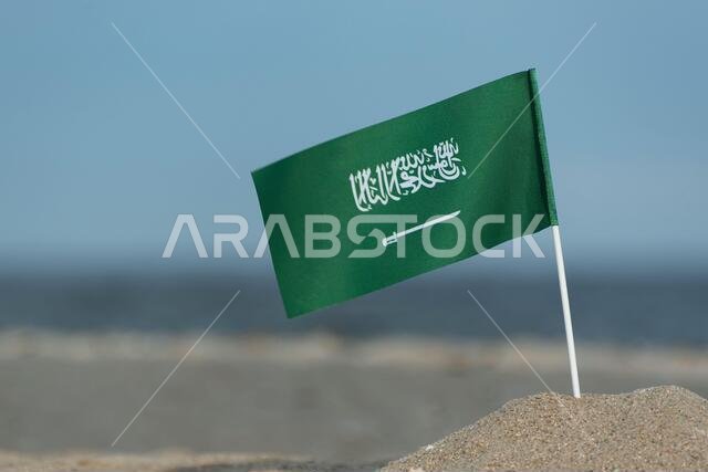 علم المملكة العربية السعودية على خلفية بحرية. علم المملكة العربية السعودية على شاطئ رملي. علم أخضر عليه سيف