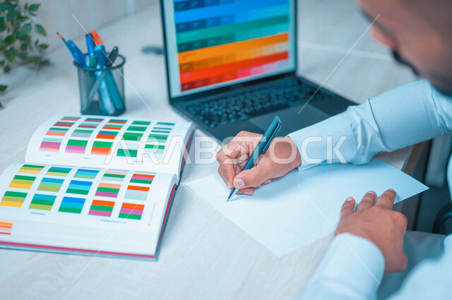 مصمم شاب عربي سعودي يقوم بتدوين الملاحظات على ورقة بيضاء , يستعين بمخطط الألوان على شاشة الكمبيوتر على مكتب لونه أبيض ، تصميم داخلي , اختيار تدرجات الألوان وتناسقها .