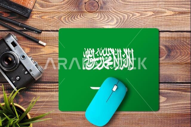 علم المملكة العربية السعودية على خلفية خشبية مع فأرة لاسلكية زرقاء على لوحة فأرة، منظر علوي. مفهوم الوسائط الرقمية.