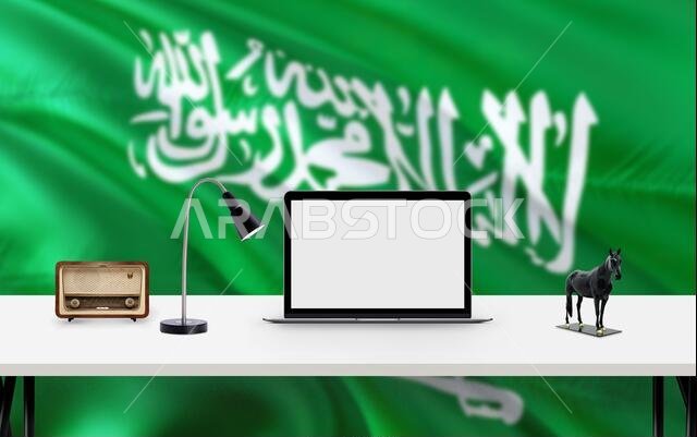 خلفية علم المملكة العربية السعودية مع مساحة عمل، وجهاز كمبيوتر مكتبي، ولوازم مكتبية على طاولة بيضاء حديثة.