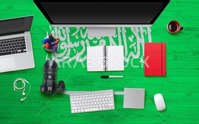خلفية علم المملكة العربية السعودية مع سماعات رأس وكاميرا ودفتر ملاحظات وفأرة على طاولة مكتب في مكتب وطني. منظر علوي مع مساحة للكتابة. صورة مسطحة.