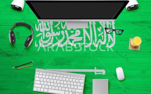 خلفية علم المملكة العربية السعودية مع سماعات رأس ولوحة مفاتيح كمبيوتر وفأرة على طاولة مكتب في مكتب وطني. منظر علوي مع مساحة للكتابة. صورة مسطحة.