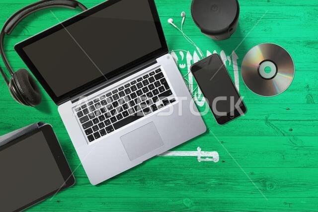 علم المملكة العربية السعودية على خلفية وطنية، طاولة خشبية. مفهوم الأجهزة المحمولة: قرص مضغوط، سماعات رأس، جهاز لوحي، حاسوب محمول. موضوع الوسائط الرقمية.