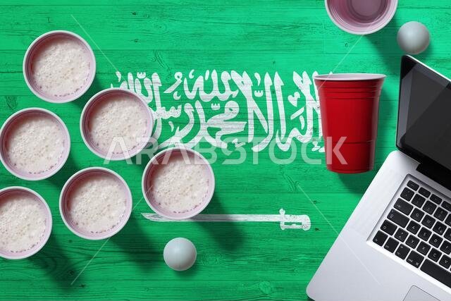Saudi Arabia flag concept with plastic beer pong cups and laptop on national wooden table, top view. Beer Pong game.