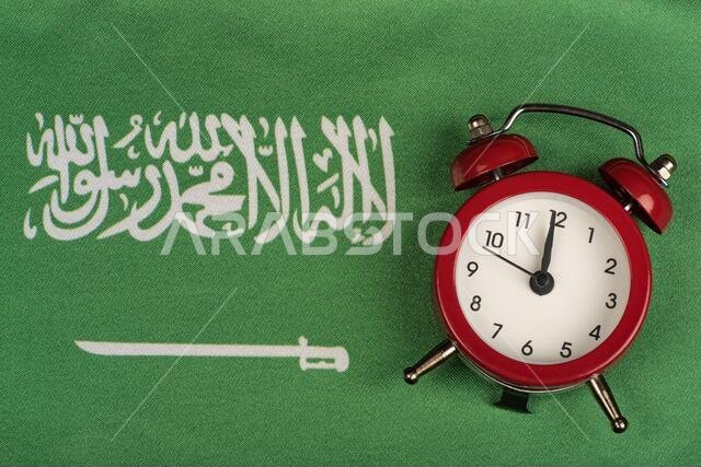 صورة مقرّبة لعلم المملكة العربية السعودية وساعة منبّهة عتيقة. علم أخضر عليه سيف.