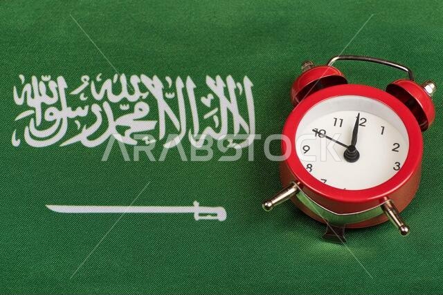 صورة مقرّبة لعلم المملكة العربية السعودية وساعة منبّهة عتيقة. علم أخضر عليه سيف.