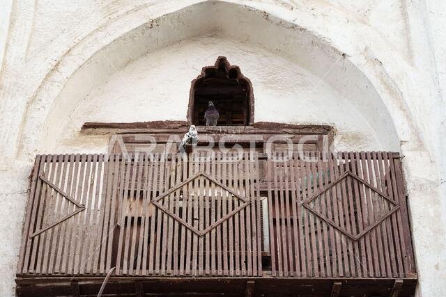 From below of pair of pigeons sitting on weathered wooden balcony of ancient building with arched window and shabby stone wall in Jeddah city in Saudi Arabia