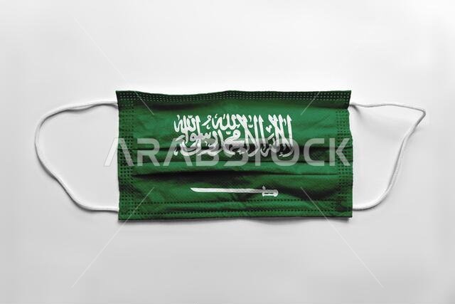 كمامة وجه مطبوع عليها علم المملكة العربية السعودية، على خلفية بيضاء، معزولة. مفهوم كوفيد-19.