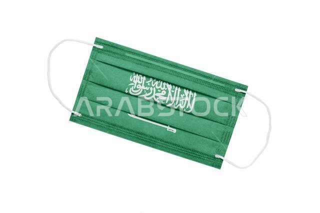 كمامة طبية عليها علم المملكة العربية السعودية، معزولة على خلفية بيضاء. مفهوم جائحة كورونا في المملكة العربية السعودية. سمة من سمات تفشي فيروس كورونا في المملكة العربية السعودية.