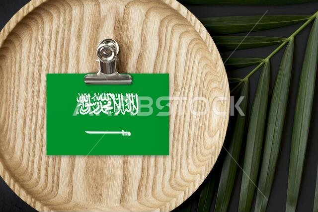 علم المملكة العربية السعودية مُلصق على لوحة خشبية. أوراق نخيل استوائية من نوع مونستيرا في الخلفية. مفهوم وطني بسيط.