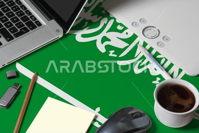 علم المملكة العربية السعودية في الأعلى، مساحة عمل مصمم مبدع مع جهاز كمبيوتر محمول، ولوحة مفاتيح، وقرص تخزين USB، وجهاز لوحي للرسم، وكوب قهوة، وفأرة.