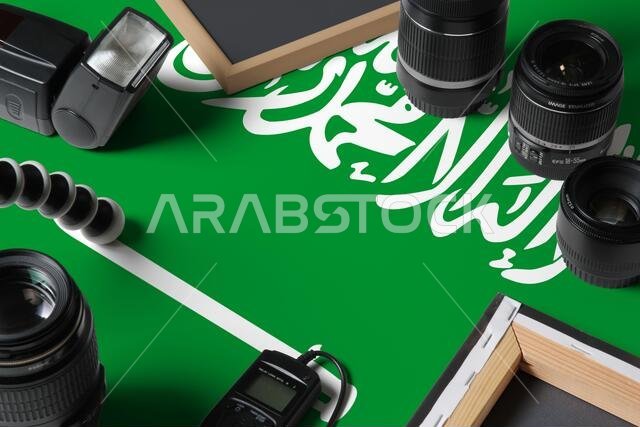 علم المملكة العربية السعودية مع صورة علوية لمعدات وأدوات المصور الشخصي على طاولة خشبية بيضاء، ومساحة للكتابة.