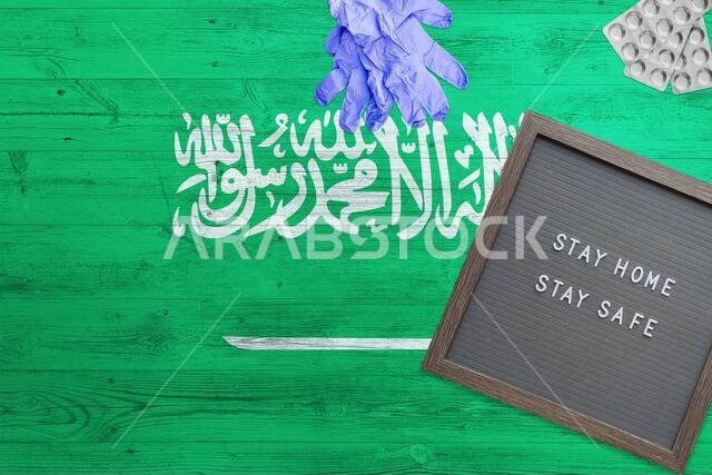 خلفية علم المملكة العربية السعودية على طاولة خشبية. لوحة كتابة "ابقَ في المنزل"، قفازات جراحية، أقراص دواء، مع رمز وطني بسيط لمفهوم كوفيد-19.